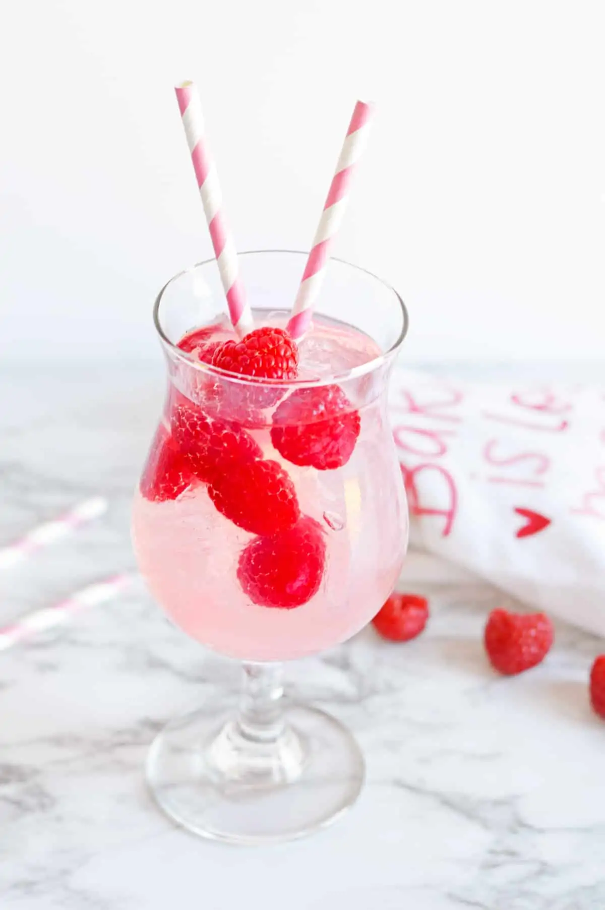 Light pink Moscato Sangria in a hurricane glass with fresh raspberries and two pink and white striped paper straws in the glass.