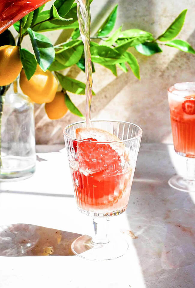 Pink grapefruit rose sangria being poured into a glass with a slice of pink grapefruit, on a bright sunny table.  Fresh lemons on branches in a vase are in the background.