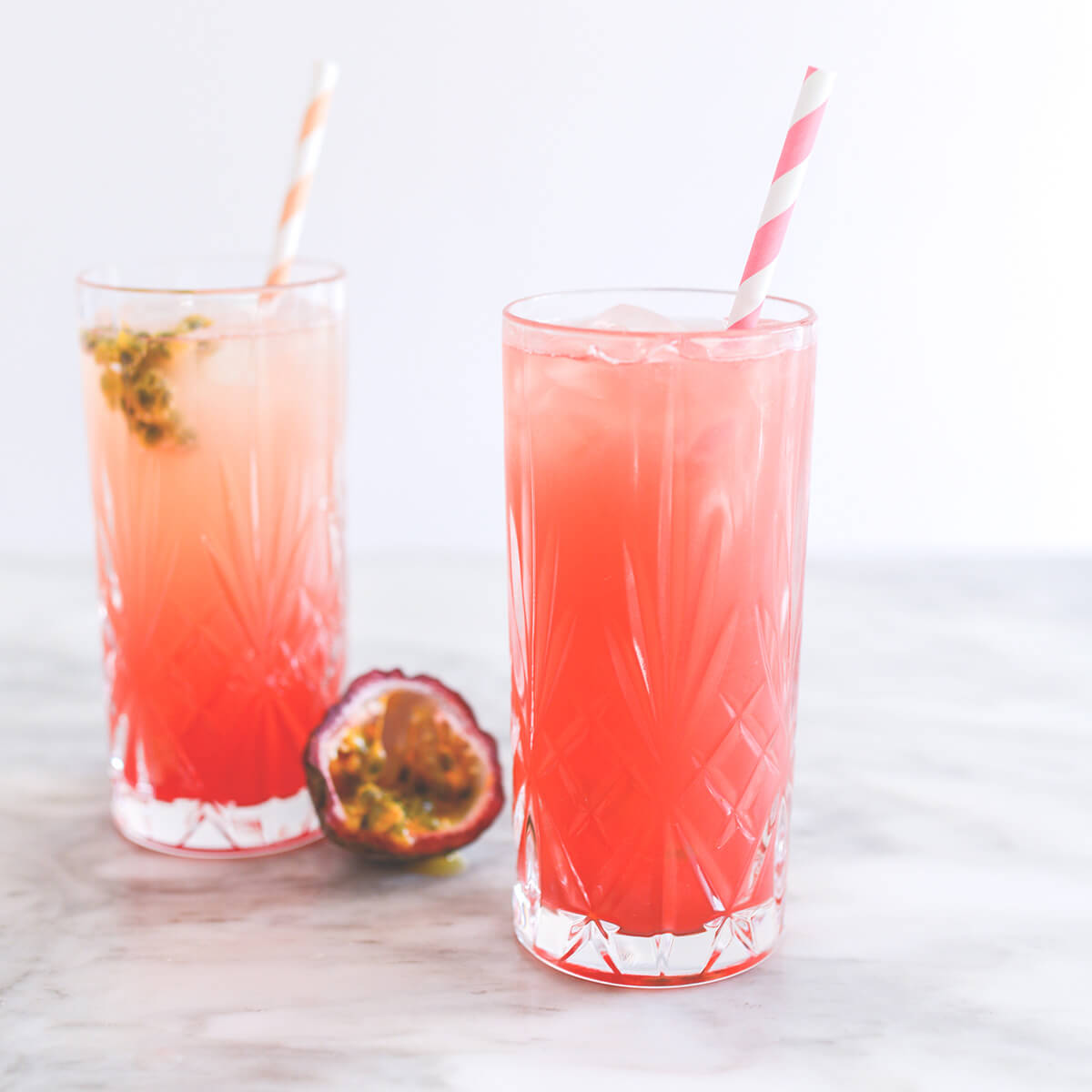 Two cut crystal highball glasses filled with passionfruit punch cocktail.  One is an ombre, layered drink ranging from deep pink at the bottom to peach at the top.  It is garnished with passionfruit seeds and half a passionfruit sits next to the glass.  The second has been stirred to create a vibrant, coral-pink cocktail.