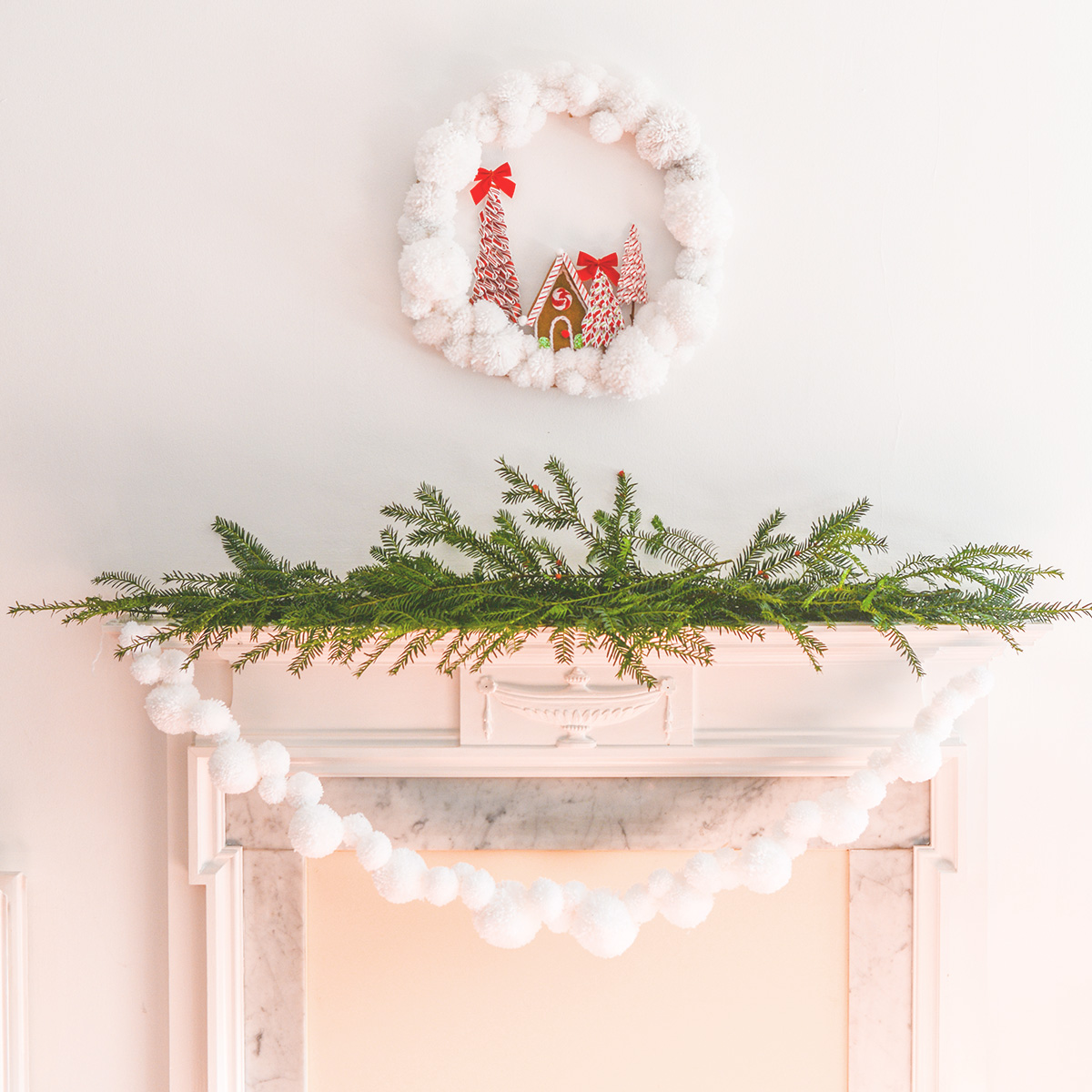 white pom pom garland hanging from a white fireplace mantel decorated with sprigs of fresh evergreen branches.  A pom pom wreath hangs above the fireplace.
