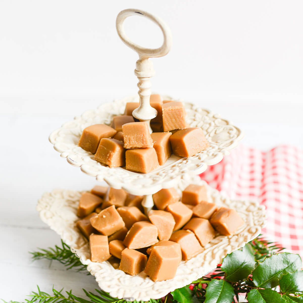 Old fashioned penuche fudge on a two tier, white serving dish.  Fresh holly and evergreen branches and a red and white checked cloth surround the tray.
