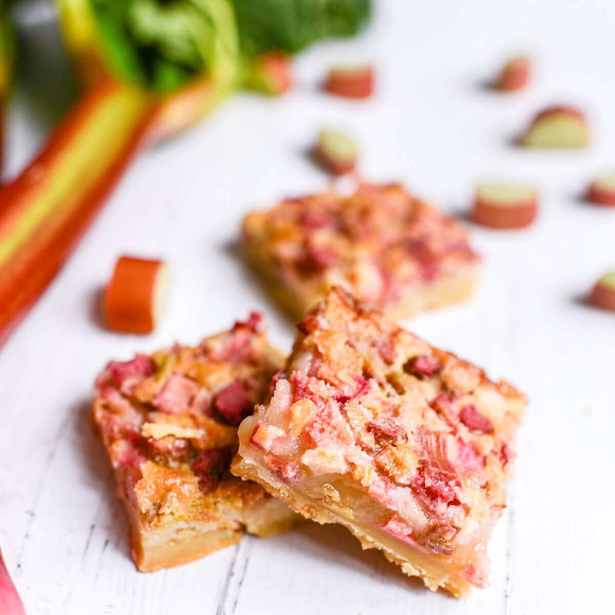 Three rhubarb custard bars cut into squares on a white wooden surface, surrounded by fresh rhubarb.