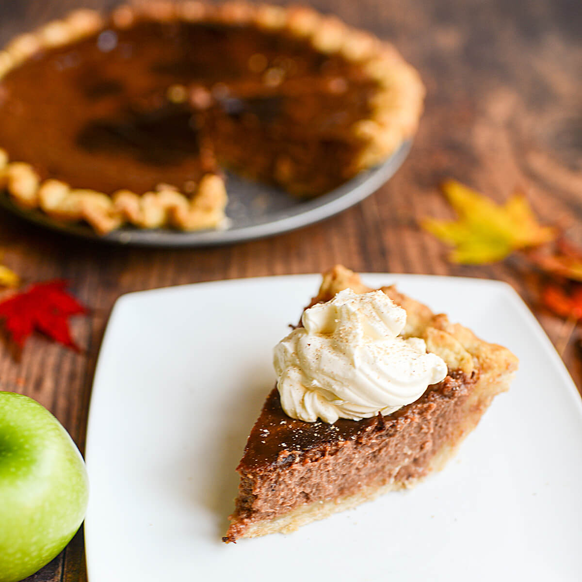 A slice of apple butter pie, topped with whipped cream, sits on a white plate in front of the pie it was cut out of.  Fall leaves and fresh apples surround the pie.