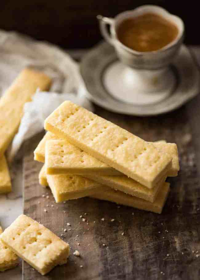 Traditional Scottish shortbread, an egg-free cookie, stacked on a wooden cutting board in front of a cup of tea on a saucer..