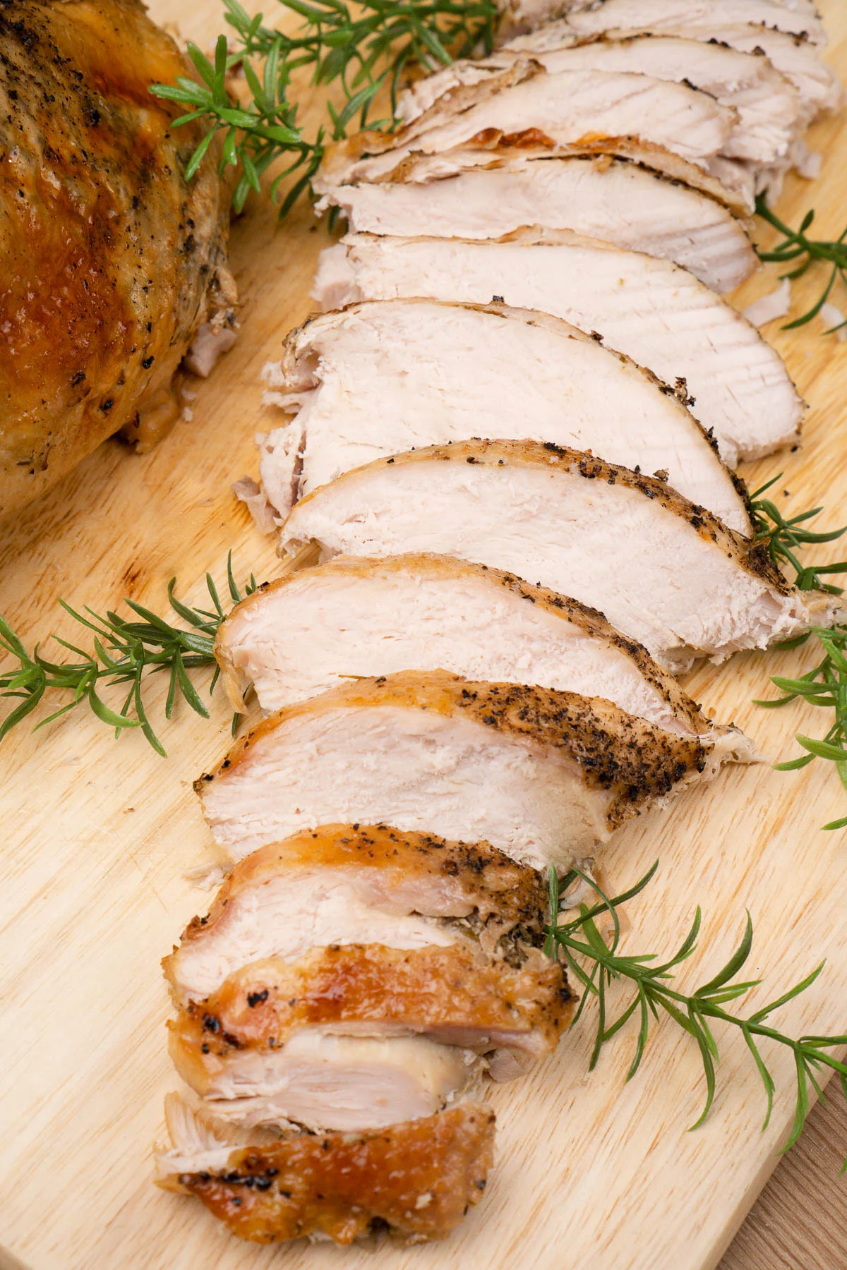 Turkey breast cooked in a crock pot, cut in slices on a cutting board, surrounded by fresh rosemary.