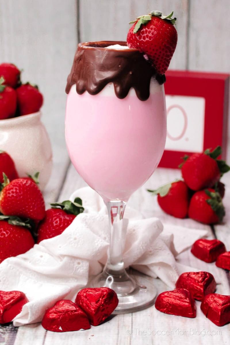 Pink frozen strawberry cocktail in a tall stemmed glass, with chocolate drip around the edge of the glass and garnished with a whole strawberry.  Fresh strawberries and heart shaped chocolates in red foil surround the drink.