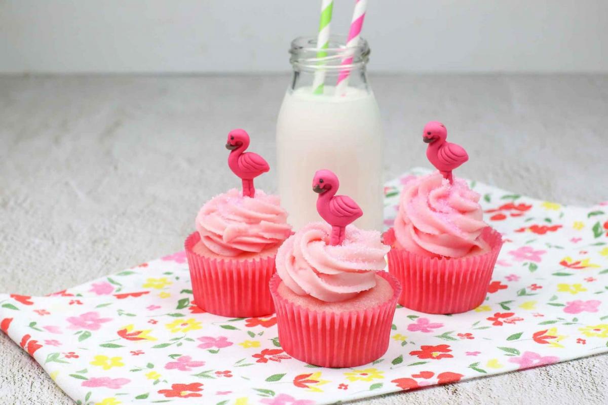 Three strawberry milk cupcakes topped with pink flamingo cupcake toppers next to a bottle of milk.