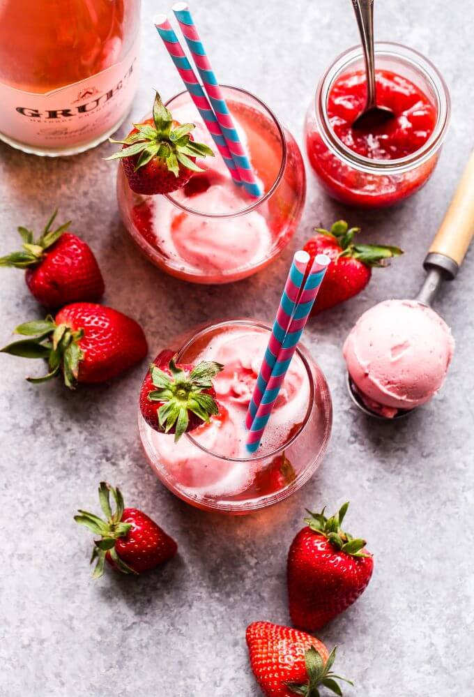 Two wine glasses filled with Strawberry Sorbet and Rose Champagne floats.  Fresh strawberries,  strawberry sorbet on an ice cream scoop, and a pot of strawberry jam surround the two glasses.
