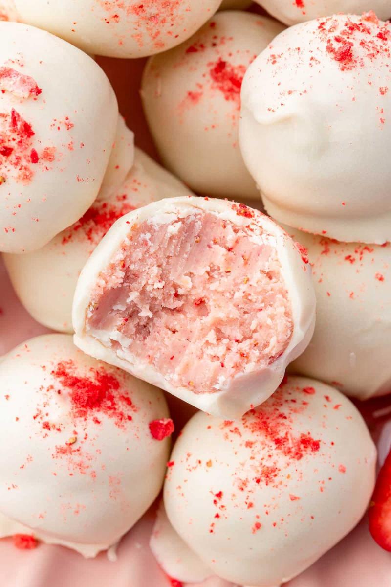 Closeup picture of a pile of white chocolate covered strawberry truffles.  The center truffle is cut in half so you can see the strawberry flavored interior of the Valentine's day candy.
