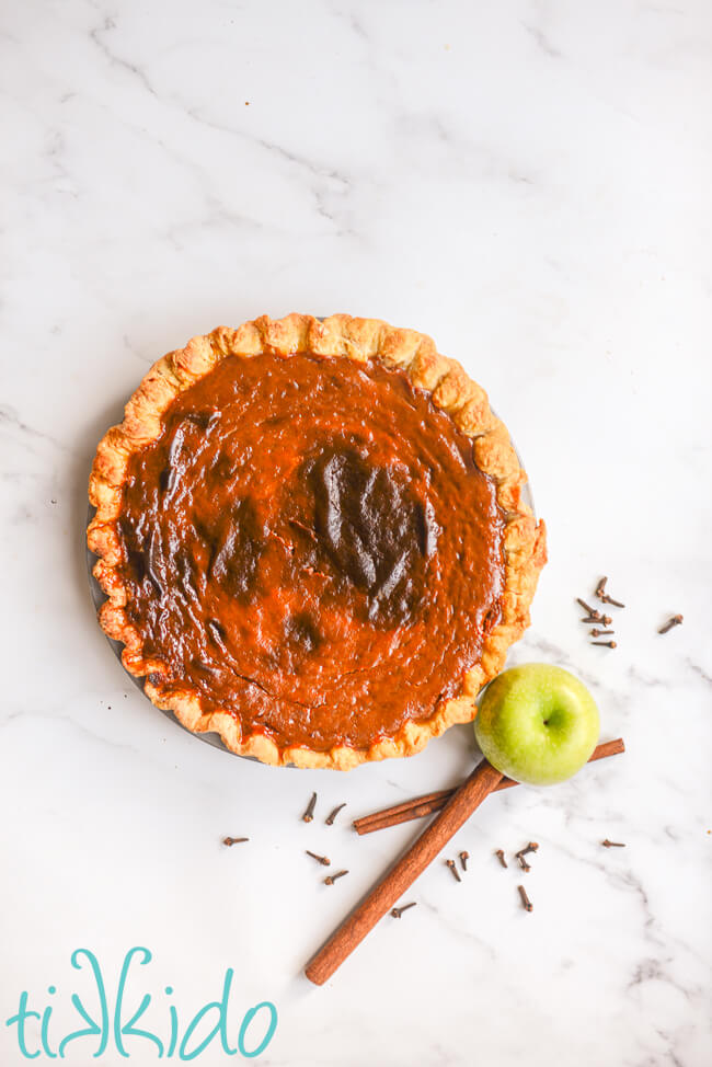 Freshly baked apple butter pie on a white marble surface, next to a fresh apple, cinnamon sticks, and whole cloves.