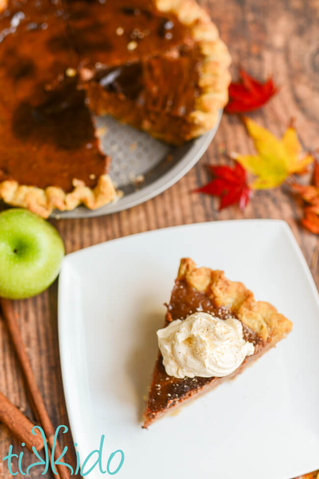 A slice of apple butter pie, topped with whipped cream and freshly grated nutmeg, on a white plate on a wooden table, surrounded by fall leaves, and apple, cinnamon sticks, and the rest of the pie.