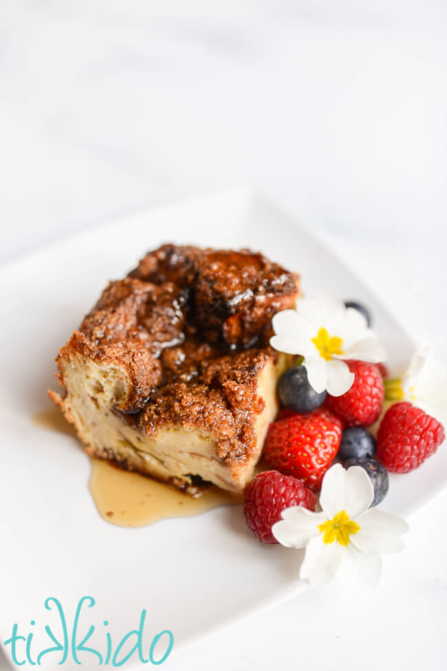 Slice of baked Brioche French toast casserole on a white plate, drizzled with maple syrup, and next to fresh berries and edible flowers.