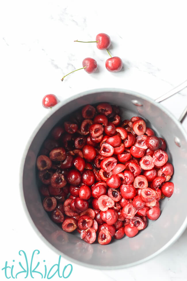 Sweet cherries, halved, pitted, and in a large saucepan on a white marble surface.
