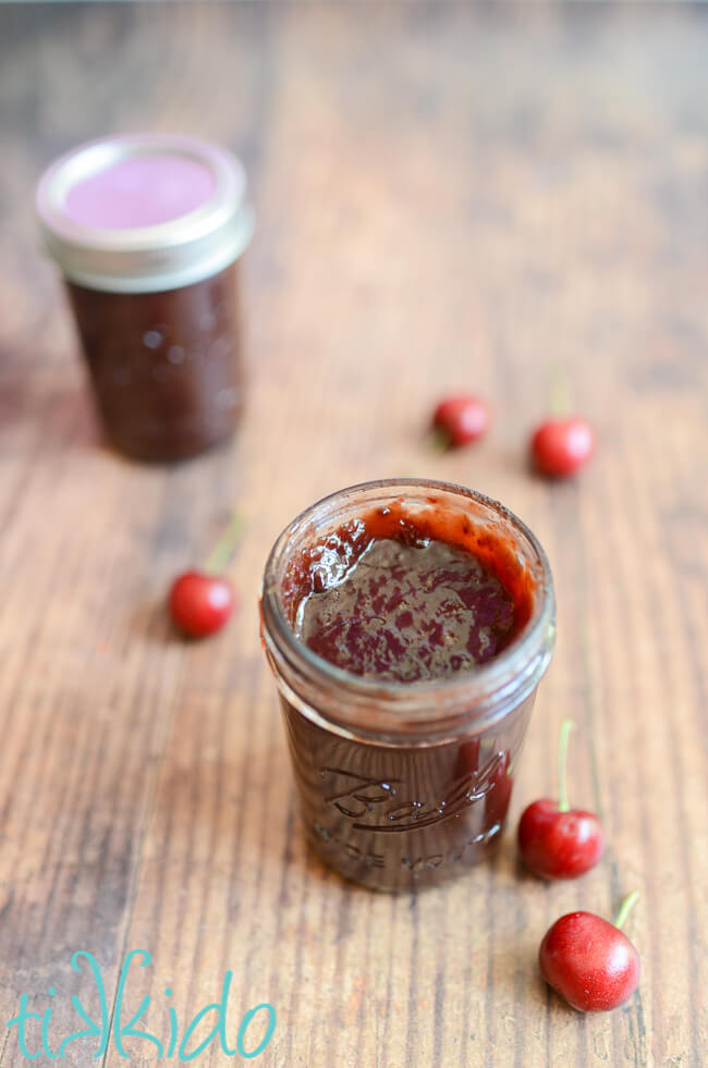One open and one closed jar of Cherry BBQ Sauce on a wooden table, surrounded by fresh cherries.