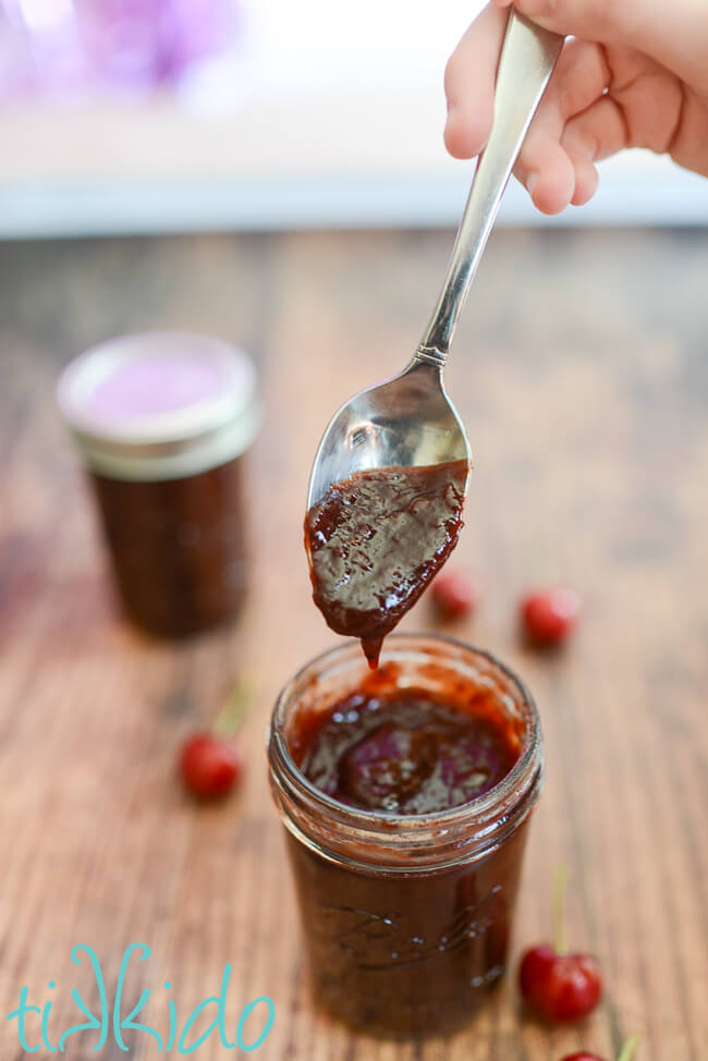 Spoon dipped in Cherry BBQ Sauce, dripping the sauce into a jar full of the cherry barbecue sauce.  Fresh cherries and another jar of the sauce surround it on a wooden table.