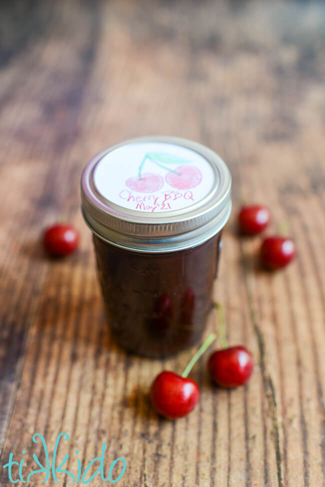 Cherry BBQ Sauce in a canning jar on a wooden table, with fresh cherries scattered around the jar.