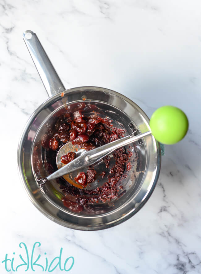 Cherry barbecue sauce ingredients being run through a food mill on a white marble surface.