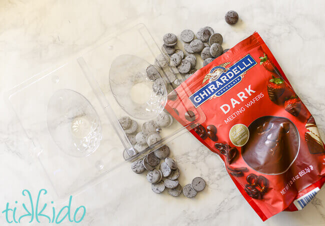 Clear plastic Easter Egg chocolate mold next to bag of Ghirardelli dark chocolate melting wafers, with discs of the melting wafers spilling on the marble surface  