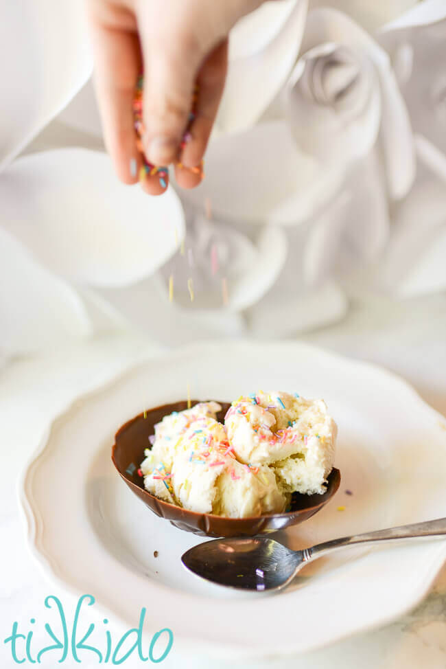 Chocolate easter egg edible ice cream bowl filled with vanilla ice cream, a hand above raining rainbow sprinkles on the ice cream.