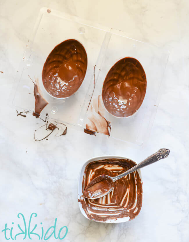 Plastic Easter egg chocolate mold coated with melted chocolate.  Empty white bowl and spoon coated in melted chocolate below.