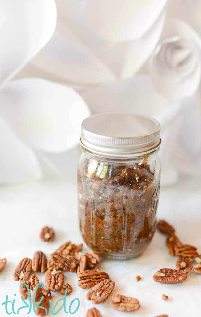 Glass ball jar filled with praline pecan ice cream topping, sitting on a white background, pecan nuts scattered around base.
