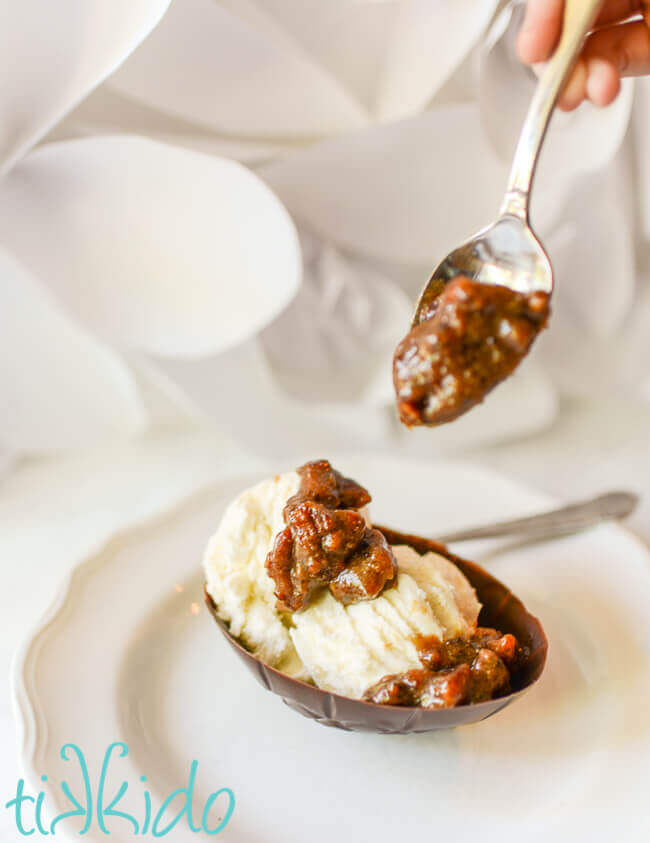 Spoon drizzling praline ice cream sauce on vanilla ice cream in an edible chocolate Easter egg bowl against a white backdrop