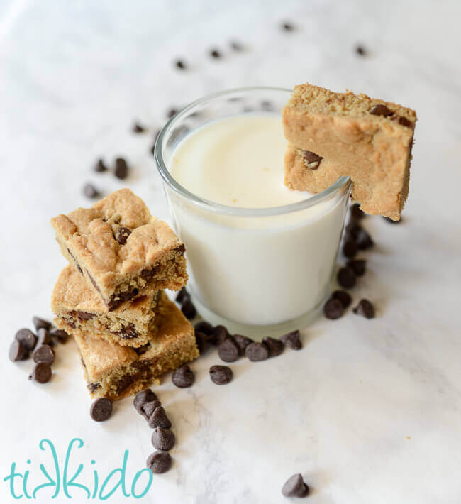 Chocolate Chip bar Cookies stacked on a white surface, next to a glass of milk with a chocolate chip cookie on the edge of the glass.
