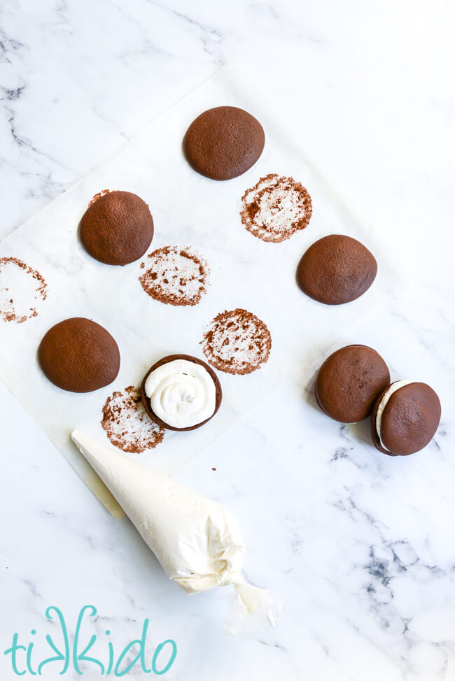 whoopie pie filling being piped into the chocolate cookies on a white marble surface.