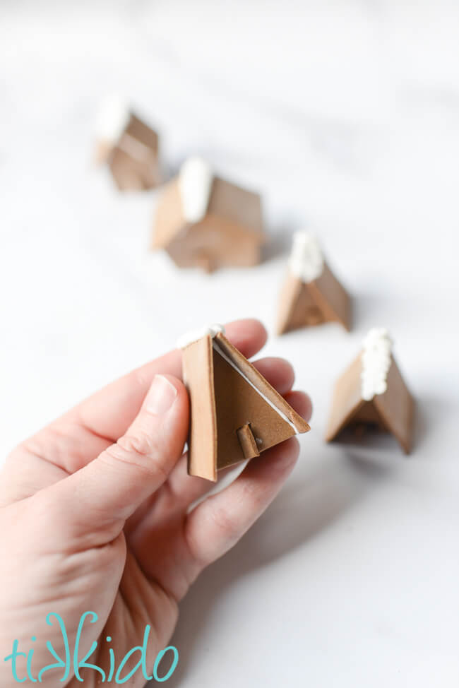 Miniature gingerbread house cupcake toppers assembled and in a line, with a hand holding one A-frame miniature gingerbread house.