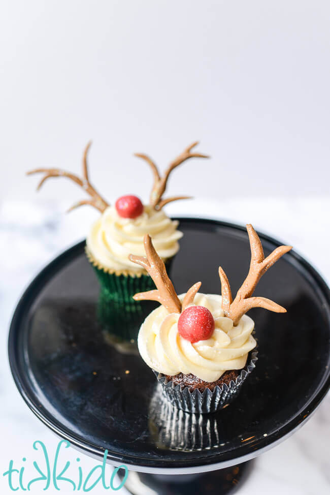 Two reindeer cupcakes on a black cake stand on a white background.