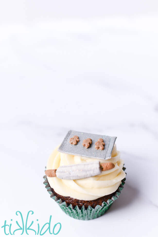 Christmas cupcake topped with a miniature gingerbread baking scene on a white marble background.