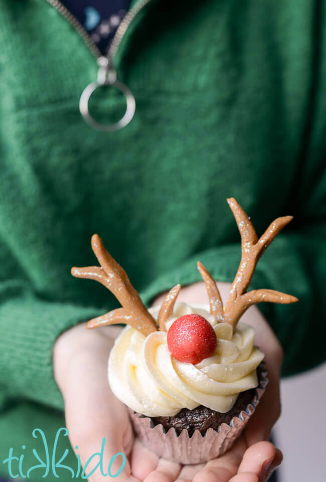 Person wearing a green sweater and holding a reindeer cupcake topped with gum paste reindeer antlers and a red Rudolph nose.