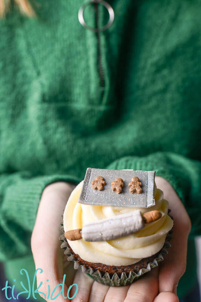 Woman in green sweater holding a cupcake topped with a miniature Christmas baking scene made out of gum paste.