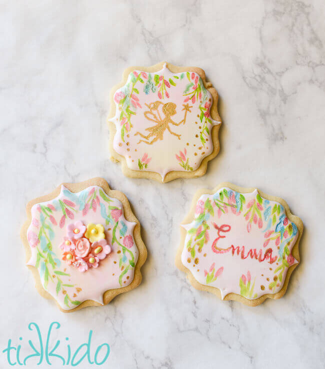 Sugar cookie covered in royal icing, with a hand painted fairy and flower design, on a white marble background.
