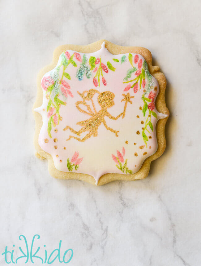 Sugar cookie covered in royal icing, with a hand painted fairy and flower design, on a white marble background.