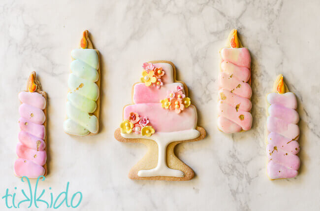 Four birthday candle shaped sugar cookies and a cookie shaped like a three tier cake on a cake stand, painted in watercolor style.