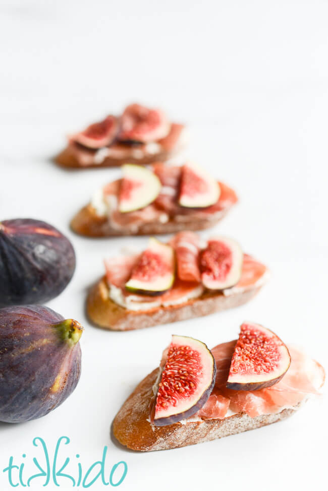 Four slices of Fig, Prosciutto, and Mascarpone Crostini next to two fresh figs on a white surface.