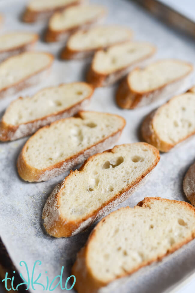 Slices of bread being toasted on a parchment lined baking sheet for making crostini.