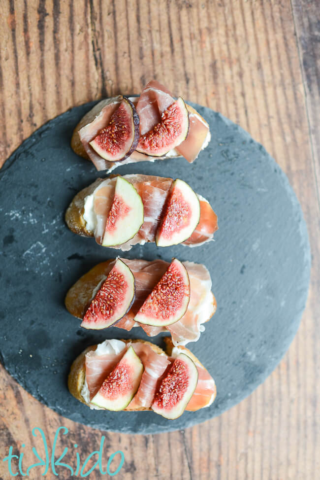 Four pieces of fig, mascarpone, and prosciutto crostini on a slate serving tray on a wooden surface.