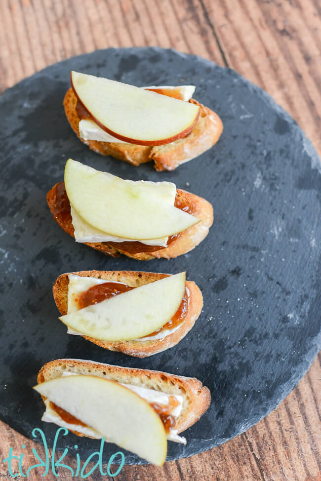 Four pieces of Apple, Brie, and Fig crostini on a slate serving tray, on a wooden surface.