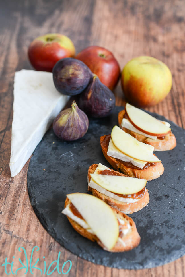 Apple, fig, and brie crostini on a slate serving tray, surrounded by a wedge of brie, fresh figs, and apples..