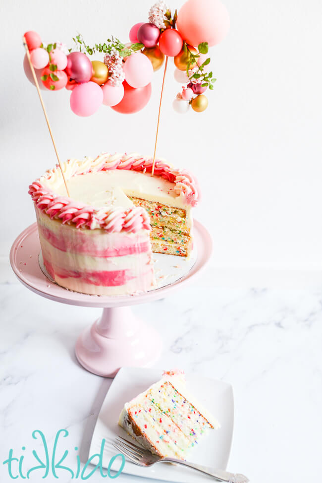 Funfetti cake decorated with pink and white icing and a mini balloon garland cake topper, on a pink cake stand, with a slice of the funfetti cake on a white plate next to it.