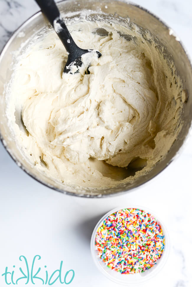 White cake batter in a mixer bowl, next to a container of sprinkles to make funfetti cake batter.