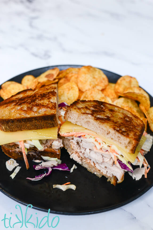 Georgia Reuben Turkey Sandwich cut in half, with one half facing the camera, so you can see the layers of turkey breast, coleslaw, and Emmental cheese.  The sandwich is surrounded by chips, and sits on a black plate.