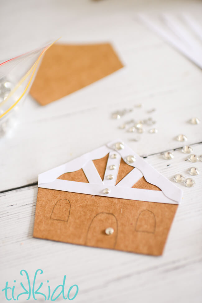 Cardboard gingerbread house shapes for a DIY Christmas garland being decorated with rhinestones.