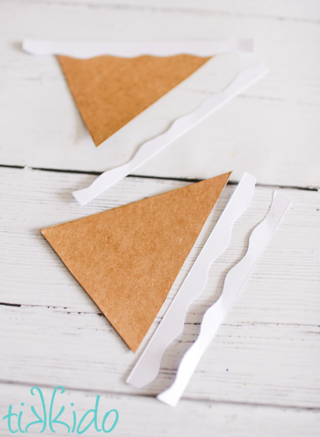 A frame gingerbread house shape cut out of cardboard, and being decorated with strips of white paper.