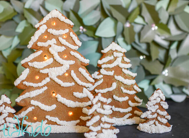 Standing gingerbread Christmas trees decorated with white royal icing and white sprinkles and lit up with LED battery operated lights.