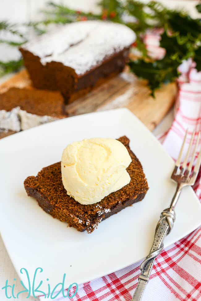 Slice of a loaf of gingerbread cake topped with a scoop of vanilla ice cream on a white plate, with the rest of the gingerbread quick bread in the background.