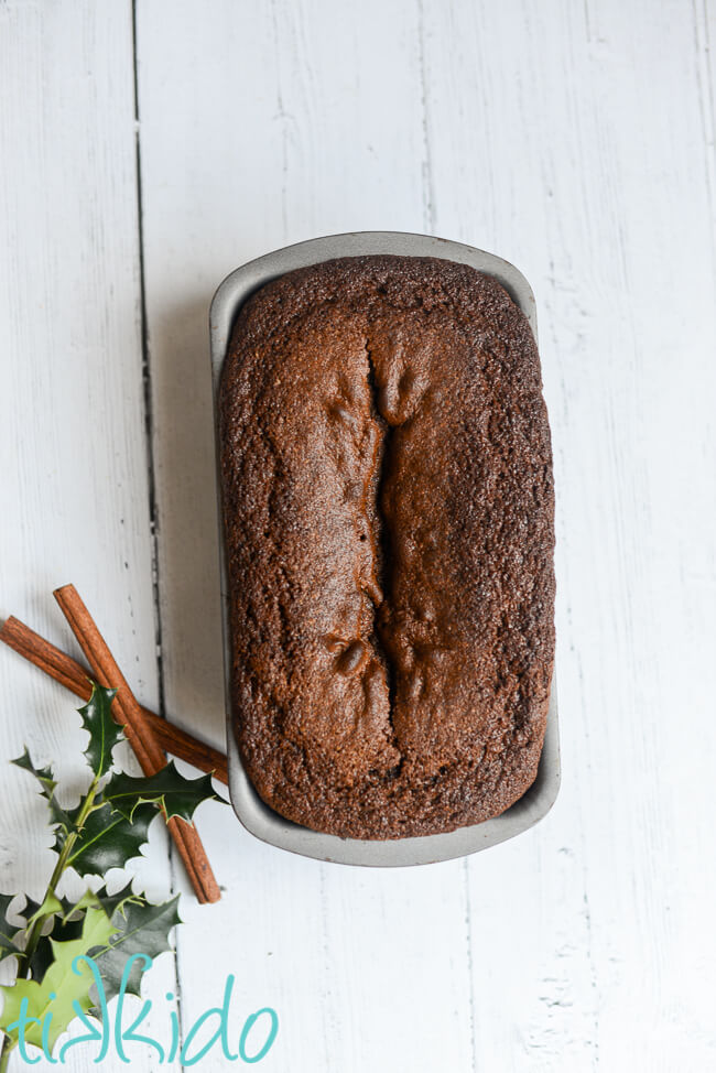 Loaf of Gingerbread Quick Bread in a bread pan on a white wooden surface, next to cinnamon sticks and holly.