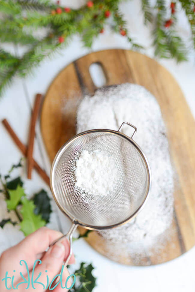 Gingerbread loaf on a wooden cutting board being dusted with powdered sugar from a small mesh strainer.